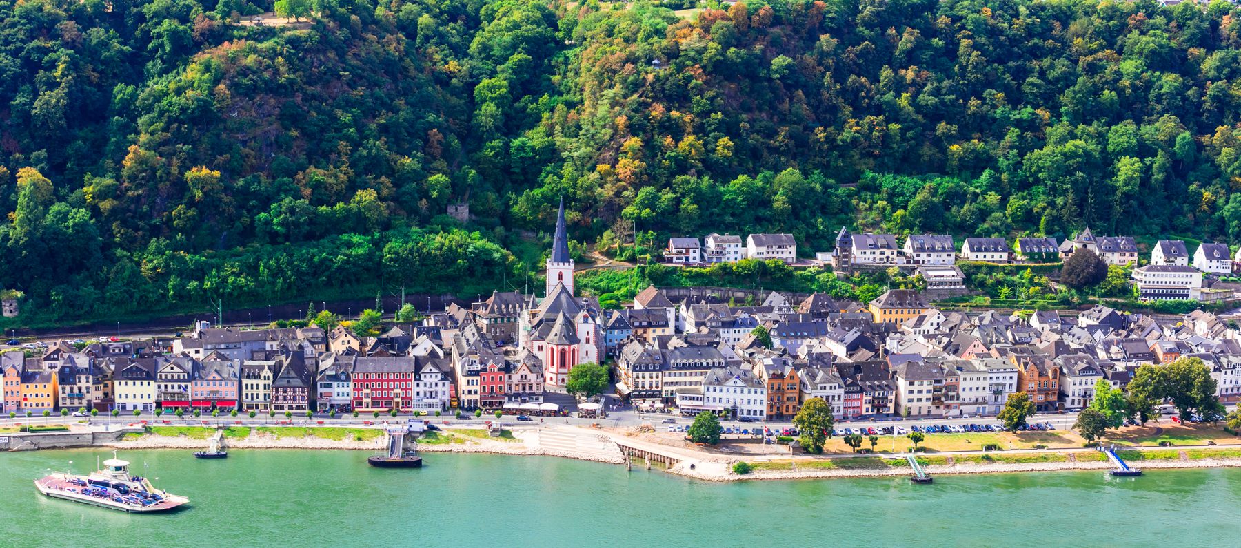 Riverside town with a church spire below wooded hills, a small boat passing